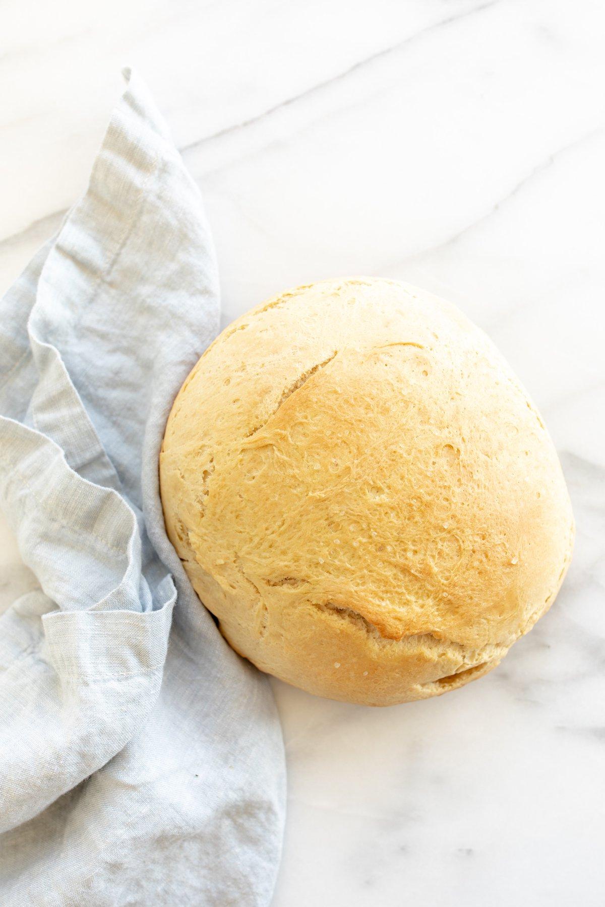 Hausgemachtes Brot im Dutch Oven 8 Eine Marmoroberfläche mit einem runden Laib hausgemachtem Brot aus dem Dutch Oven, daneben eine blaue Leinentischdecke.