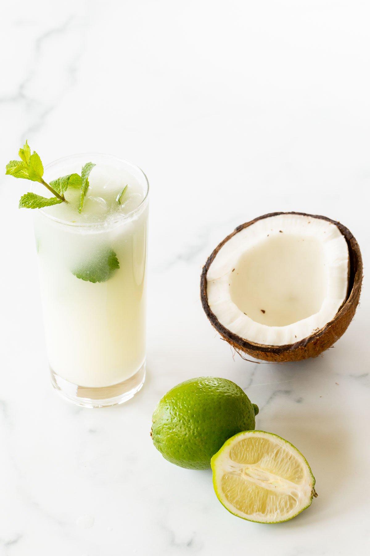 Kokosnuss-Mojito 9 Ein Kokos-Mojito, garniert mit Minze, auf einer Marmorfläche. Limetten und eine offene Kokosnuss daneben.