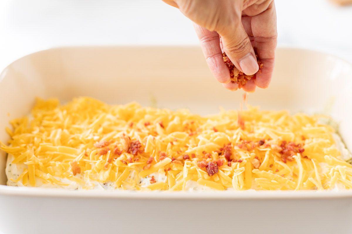 Eine Hand verteilt gehackten Speck über eine weiße Auflaufform voller 'Crack Chicken', bereit zum Backen.