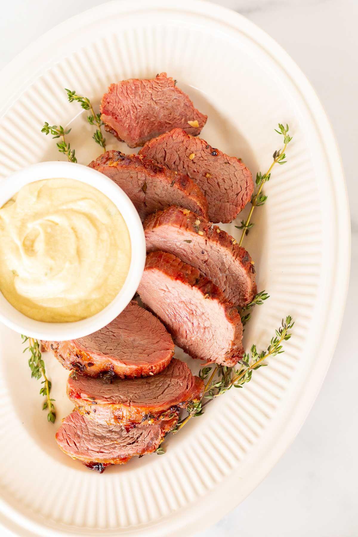 Eine weiße Schale voller Dijon-Senfsauce, umgeben von Rinderfilet auf einer Platte