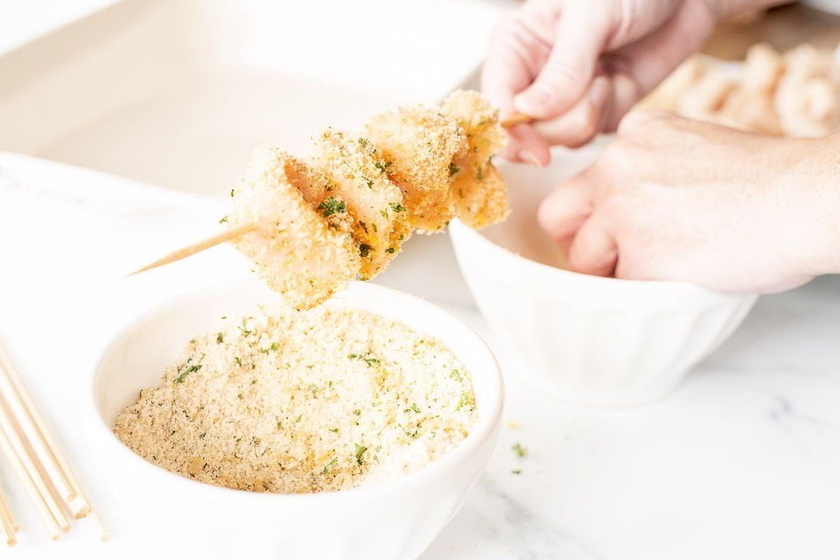 Eine Hand, die einen Hähnchen-Spiedini-Spieß über einer Schüssel mit Paniermehl hält.