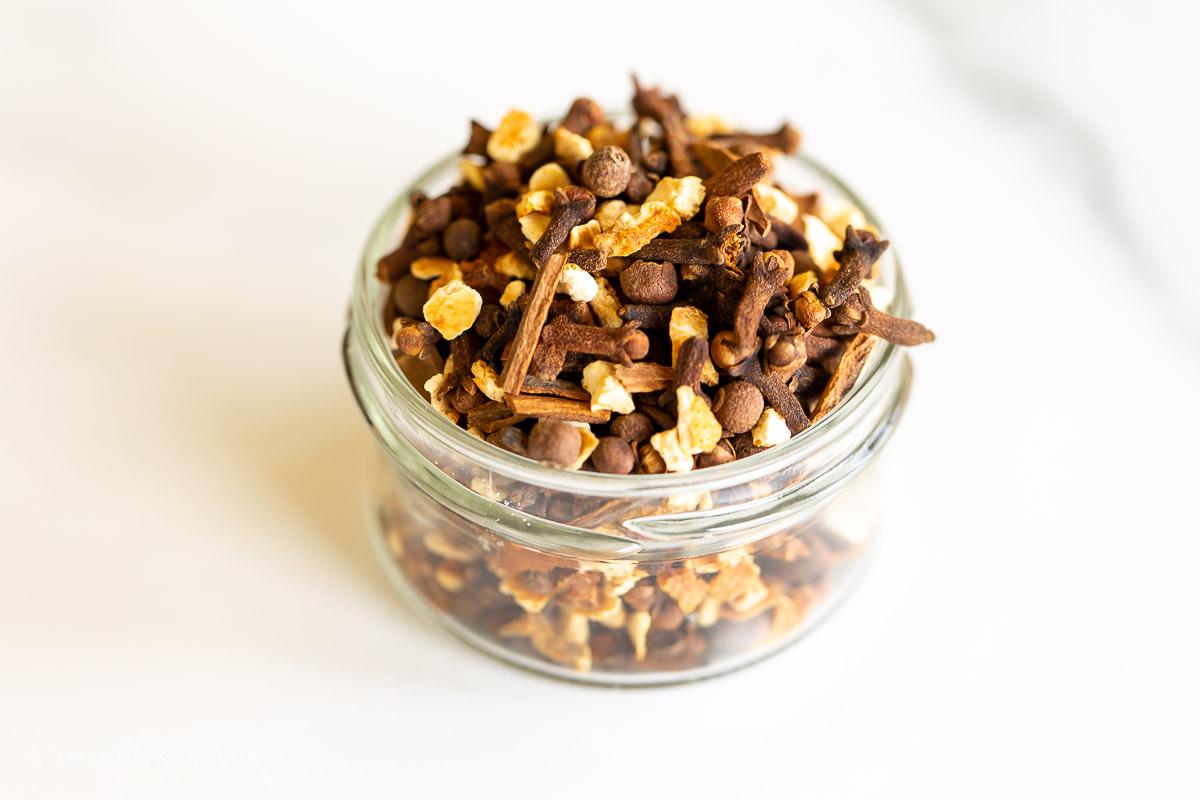 Glühweingewürz Rezept für Glühwein, gewürzten Apfelpunsch oder duftendes Potpourri, mit einer Mischung aus Zimtstangen, Piment, ganzen Nelken und Orangenschalen. Ein Glasgefäß gefüllt mit Zimt und anderen Gewürzen, die das Aroma von Glühweingewürz verströmen, auf einer Küchenfläche.