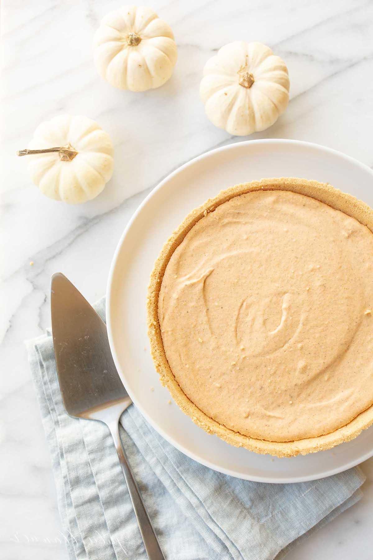 Einfaches Kürbis-Käsekuchen-Rezept ohne Backen 9 Ein No-Bake Kürbis-Käsekuchen auf einem weißen Teller, daneben ein Tortenheber und kleine weiße Kürbisse im Hintergrund.