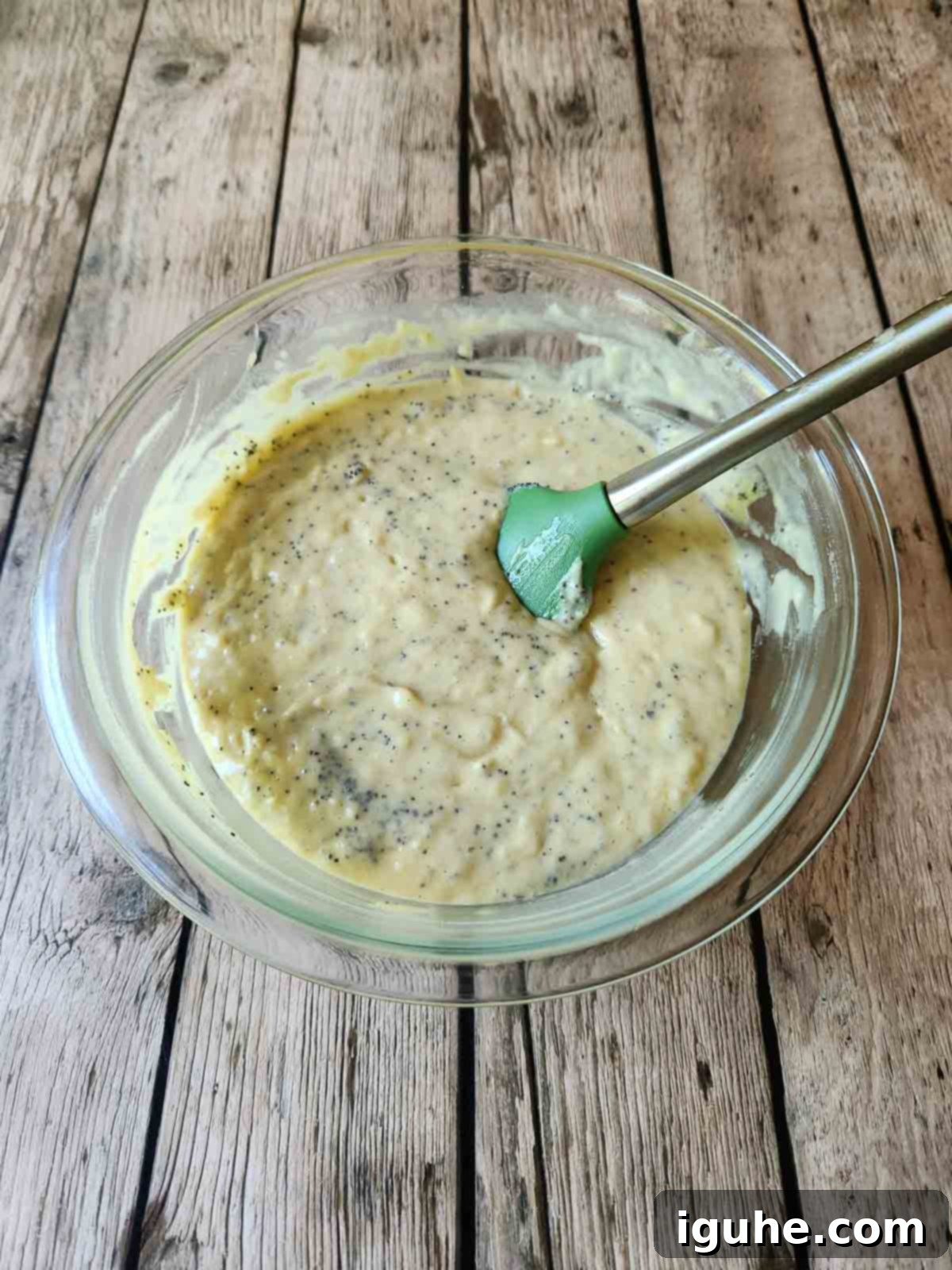 Zesty Lemon Poppy Seed Gems 5 A spatula gently folds rich, creamy muffin batter in a clear mixing bowl, ensuring all ingredients are just combined for perfect texture.