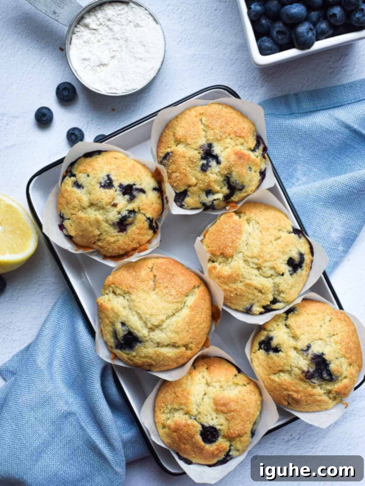 Artisan Zesty Lemon Blueberry Muffins 2 Overview image of bakery-style blueberry lemon muffins on a white background with fresh blueberries scattered next to a blue napkin.