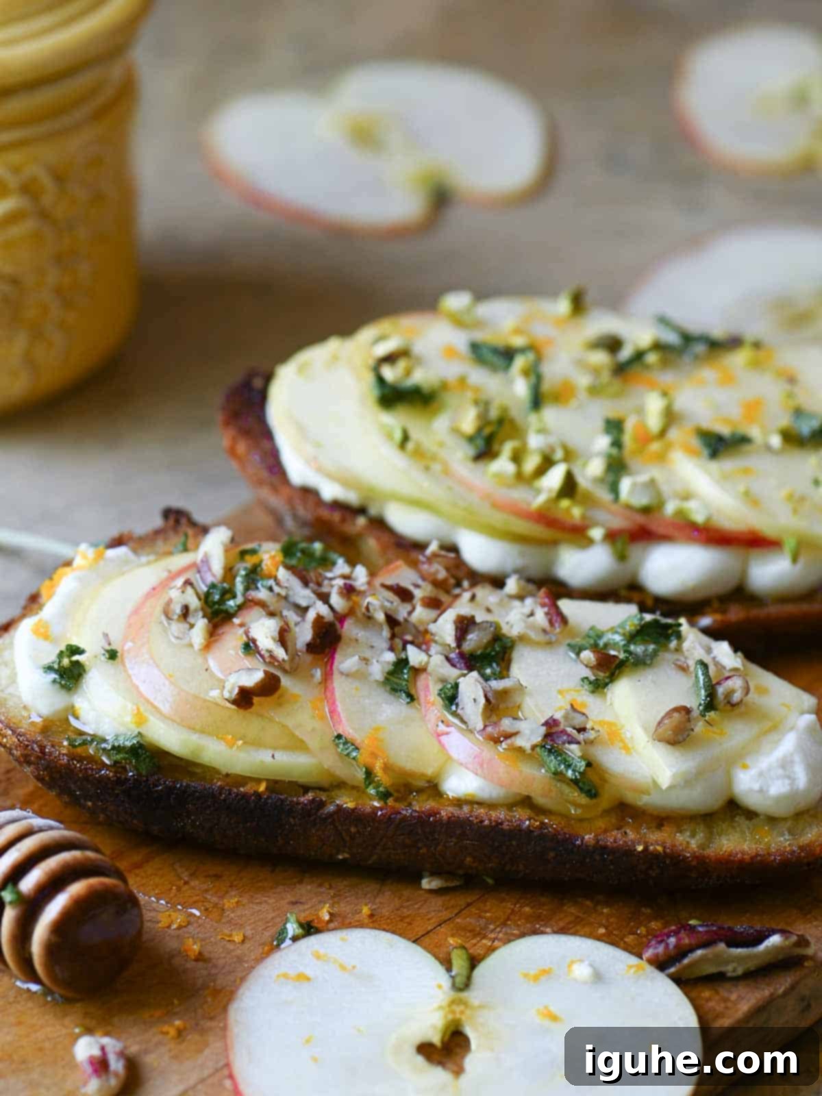 Creamy Ricotta Apple Toast with Sage-Kissed Honey 3 Two slices of apple toast on a wooden cutting board, garnished with fresh sage and nuts.