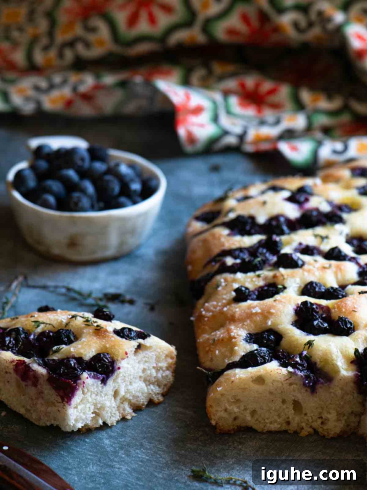 Sweet and Savory Blueberry Thyme Focaccia 2 A slice of blueberry focaccia on a grey surface with blueberries in the background.