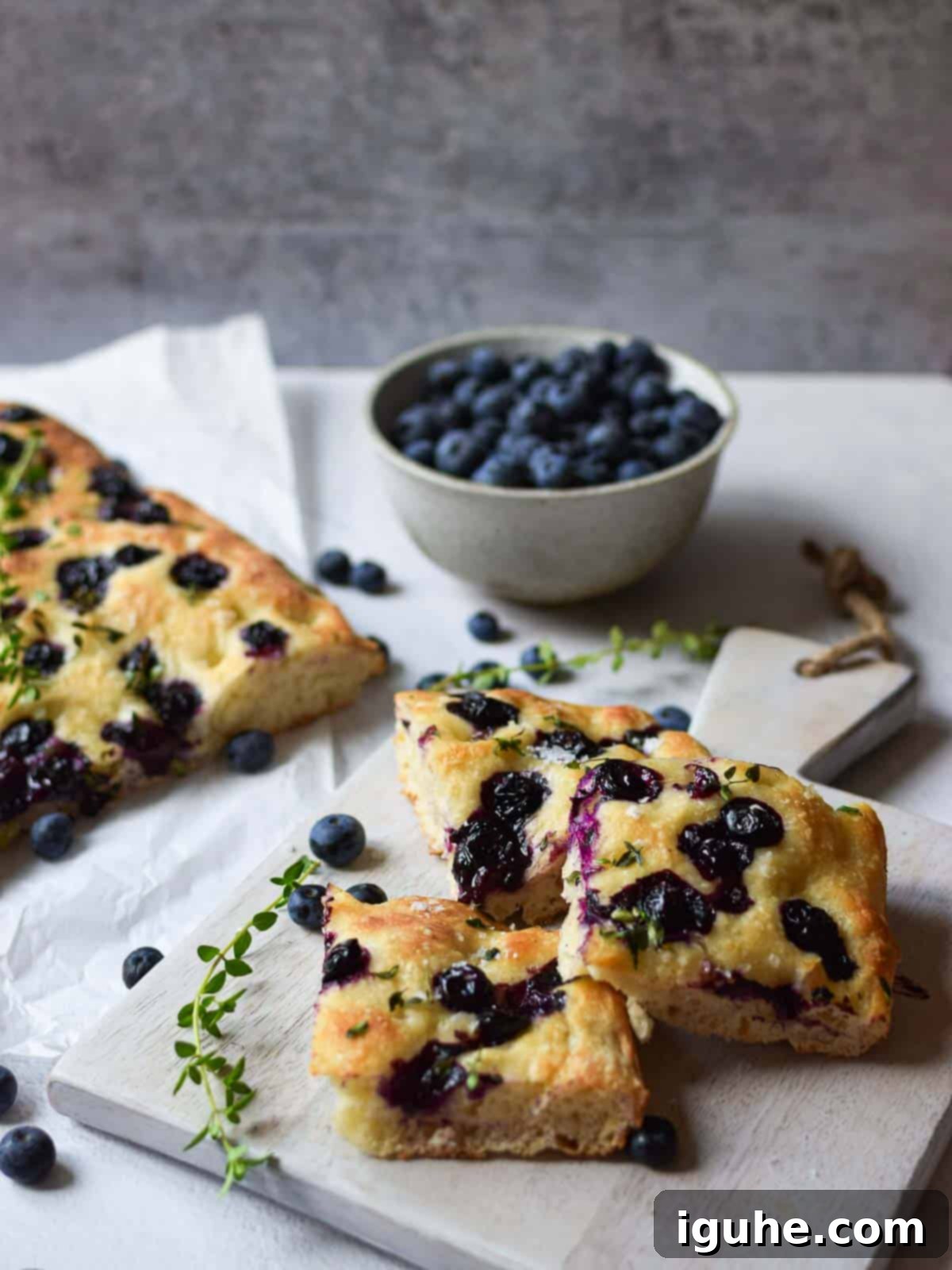 Sweet and Savory Blueberry Thyme Focaccia 3 Angled view of blueberry thyme focaccia on a grey cutting board with blueberries in the background.