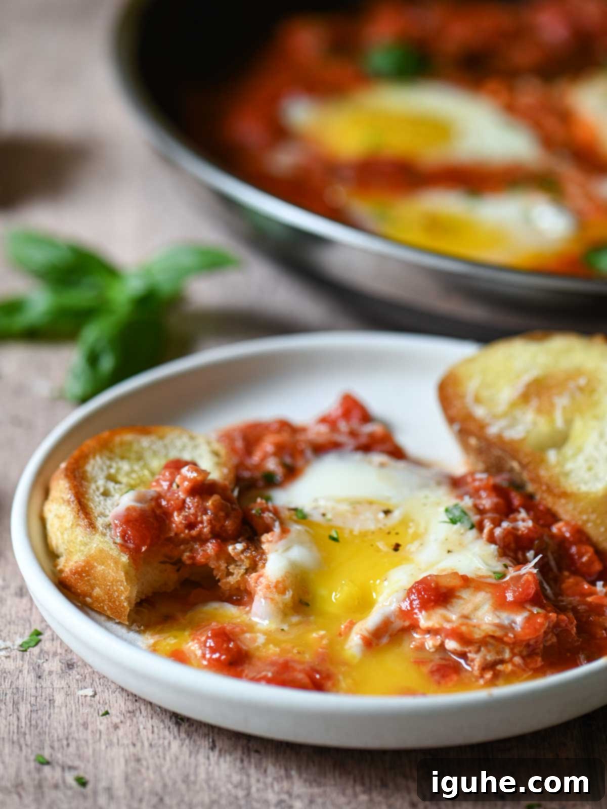 Domates Alevinde Yumurtalar 10 Savory eggs in tomato sauce with garlic bread on a white plate, garnished with fresh basil.