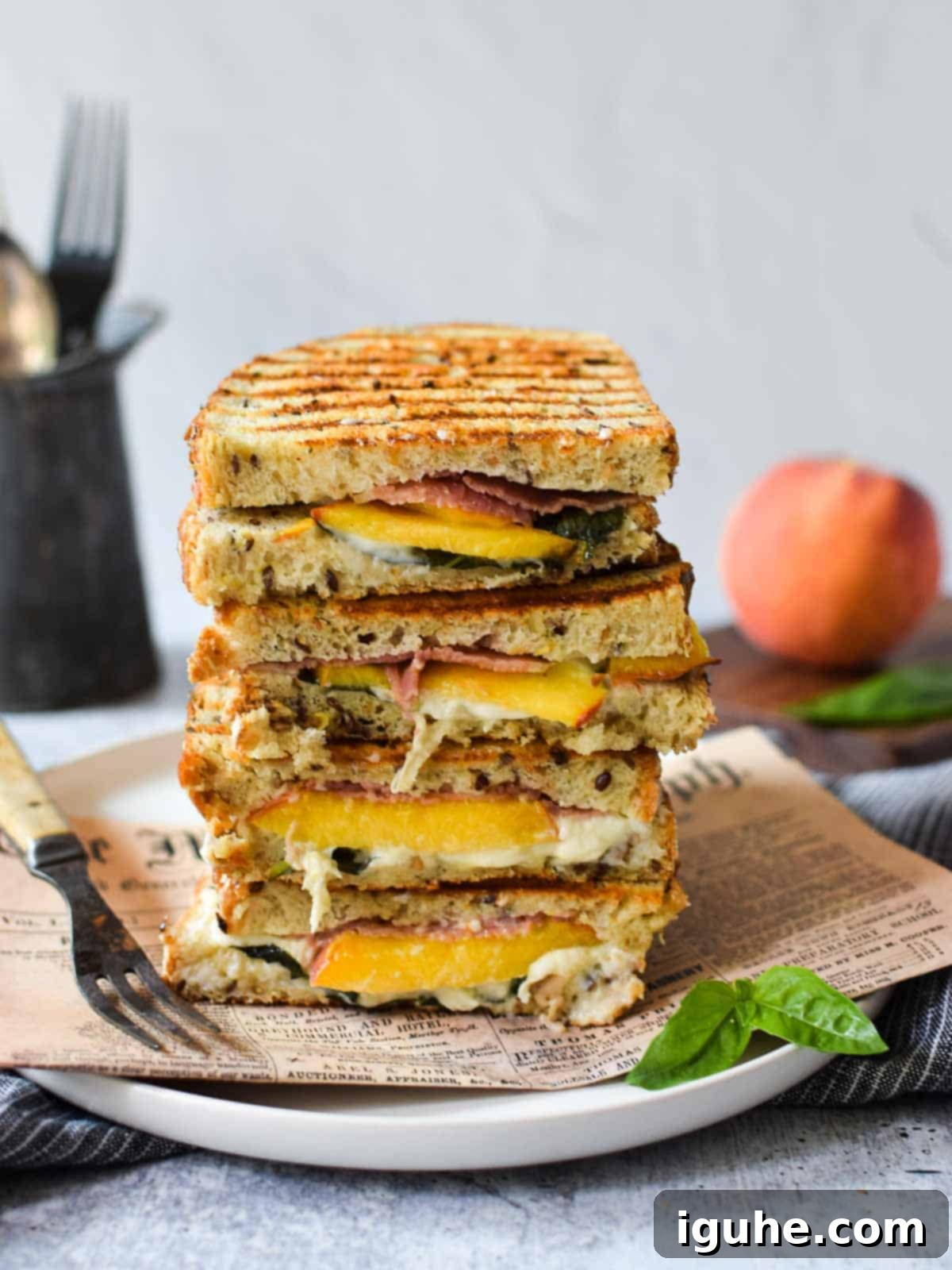 Sweet and Savory Peach Prosciutto Burrata Panini 7 An eye-level view of two peach, prosciutto, and burrata paninis stacked on a white plate, with a fresh peach in the background.