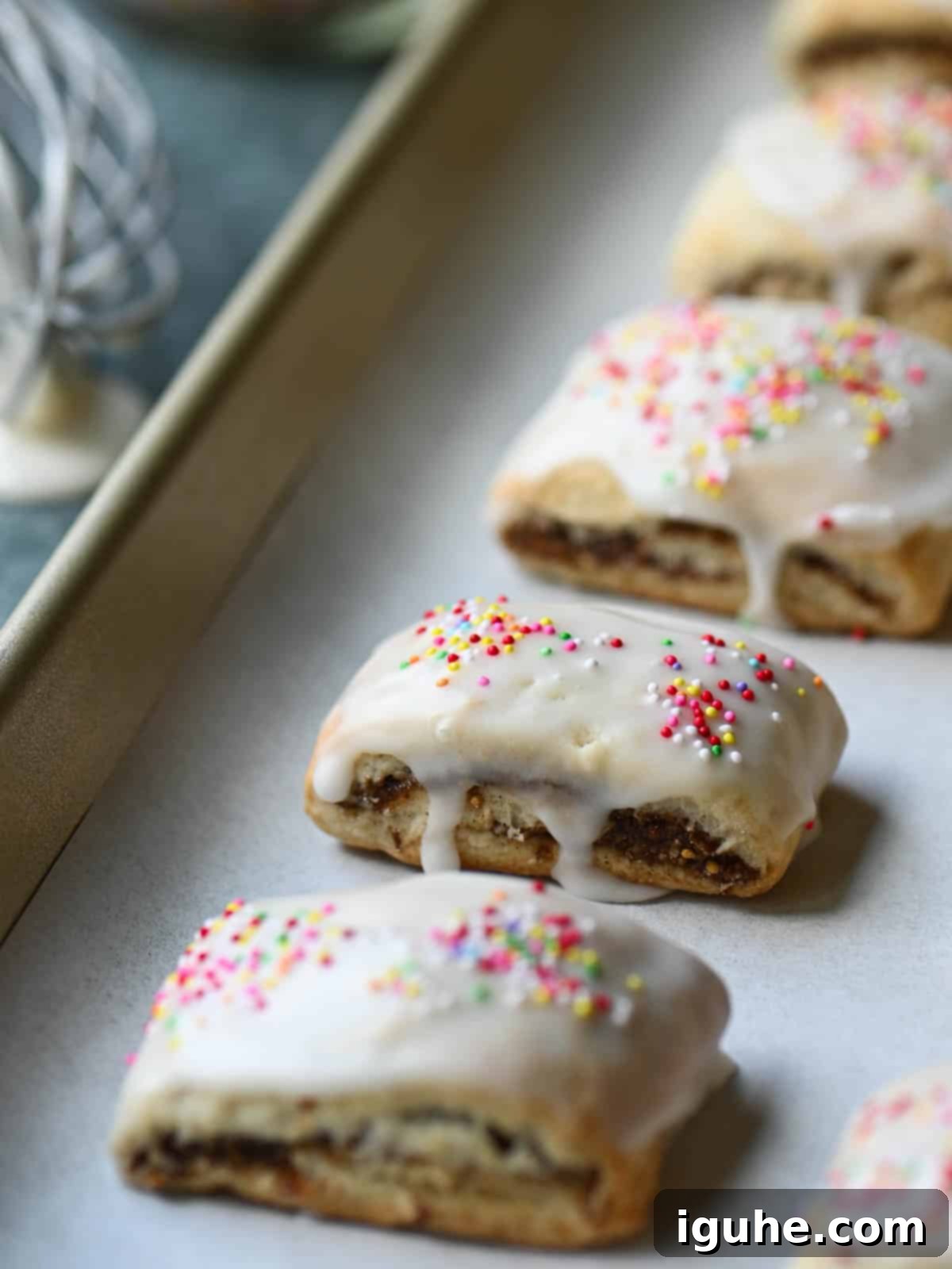 Sicilian Cuccidati Festive Fig Cookies 2 Iced fig cookies on a baking tray with white parchment paper, glistening with glaze and colorful sprinkles.
