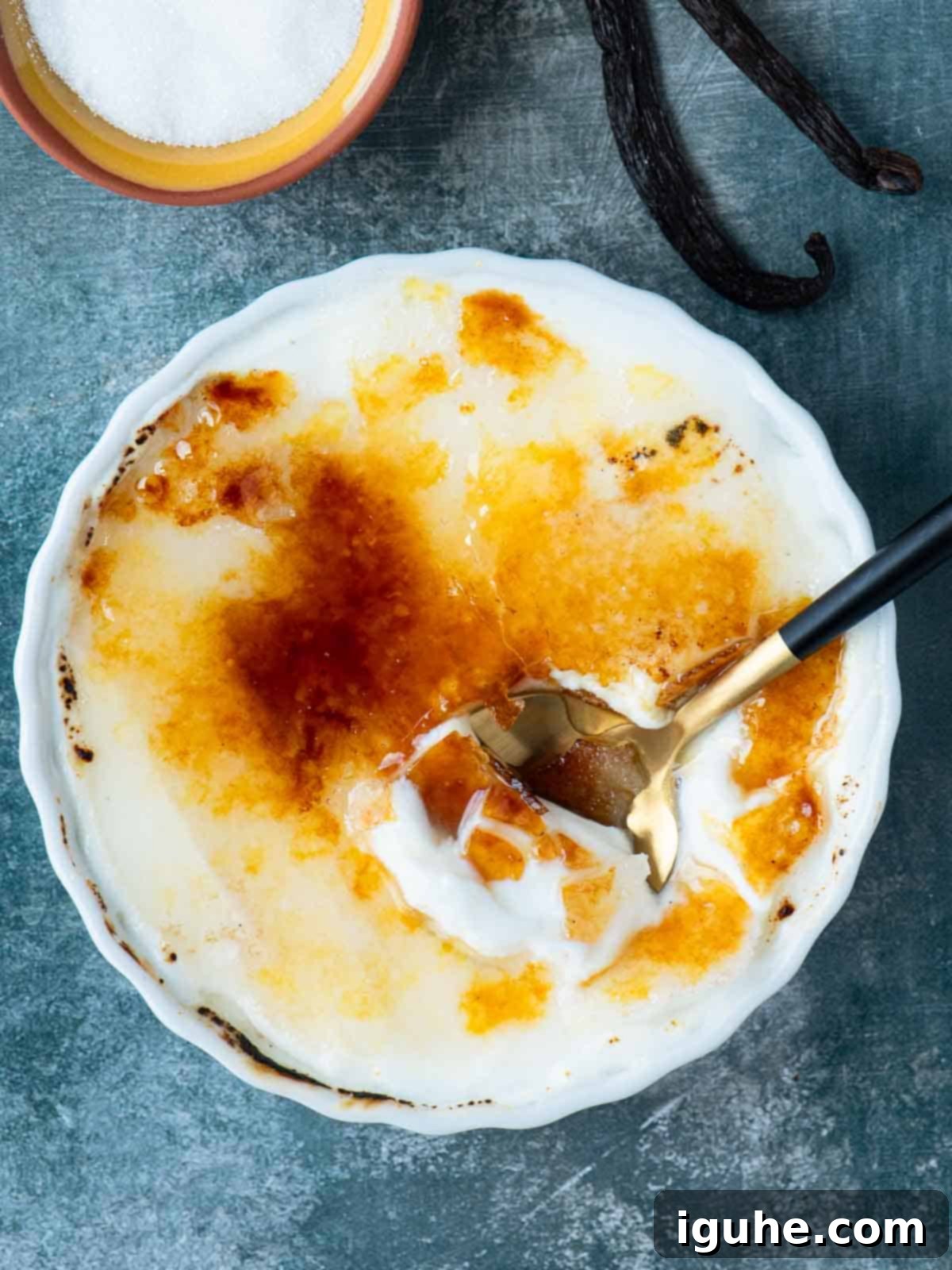 Effortless Vanilla Yogurt Brulee 6 Overhead view of creme brulee on a blue surface with a spoon dipped into it.