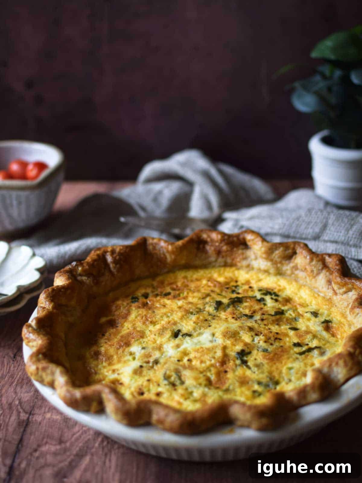 Angled view of tomato, leek and feta quiche on a brown surface with tomatoes and a linen in the background