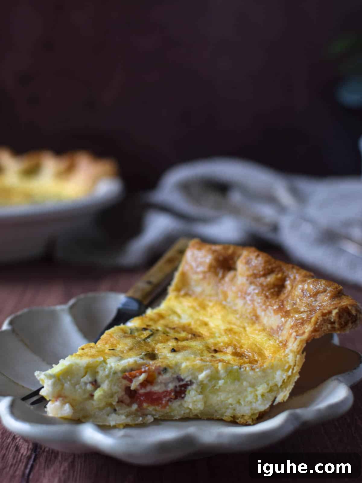 Sun-Kissed Tomato Feta Leek Quiche 8 Slice of tomato, leek and feta quiche on a brown cutting board with quiche in the background