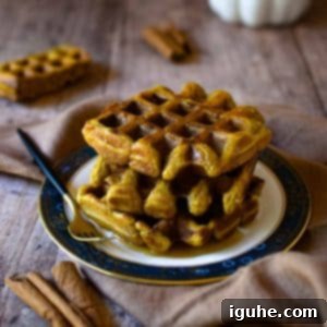 Pumpkin Spice Belgian Waffles 7 Pumpkin waffles stacked on a plate with maple syrup and cinnamon on a wooden surface.