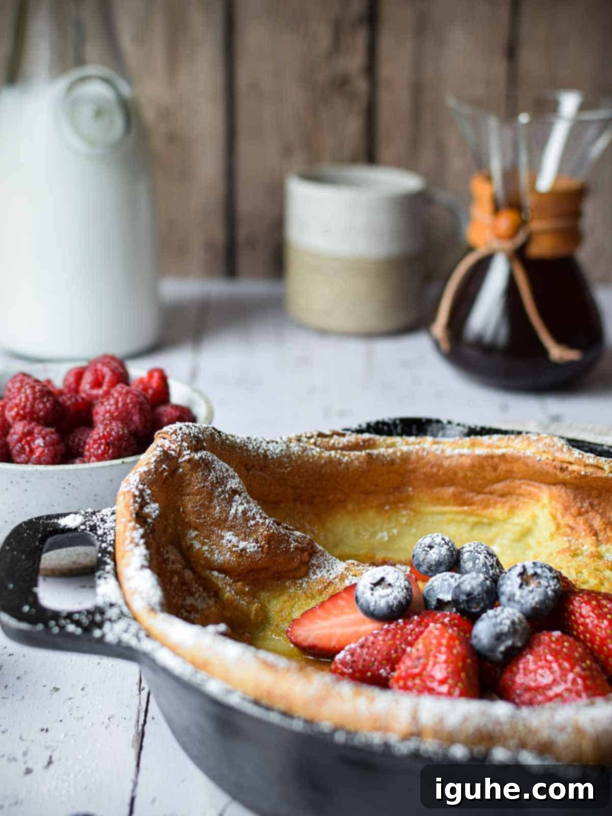 Vanilla Cloud Dutch Baby Pancake 3 Eye-level view of a Dutch baby in a cast iron skillet with fruit and coffee in the background.