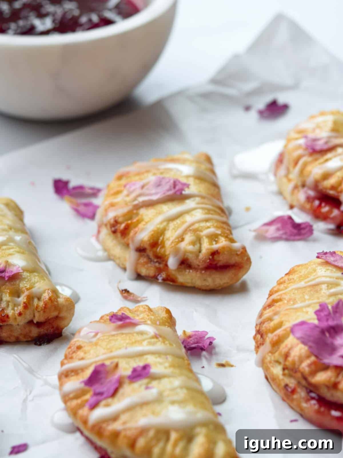 Close-up of freshly baked strawberry mini pies on white parchment paper, with a jar of strawberry jam and fresh strawberries in the blurred background, highlighting their vibrant color and inviting texture.