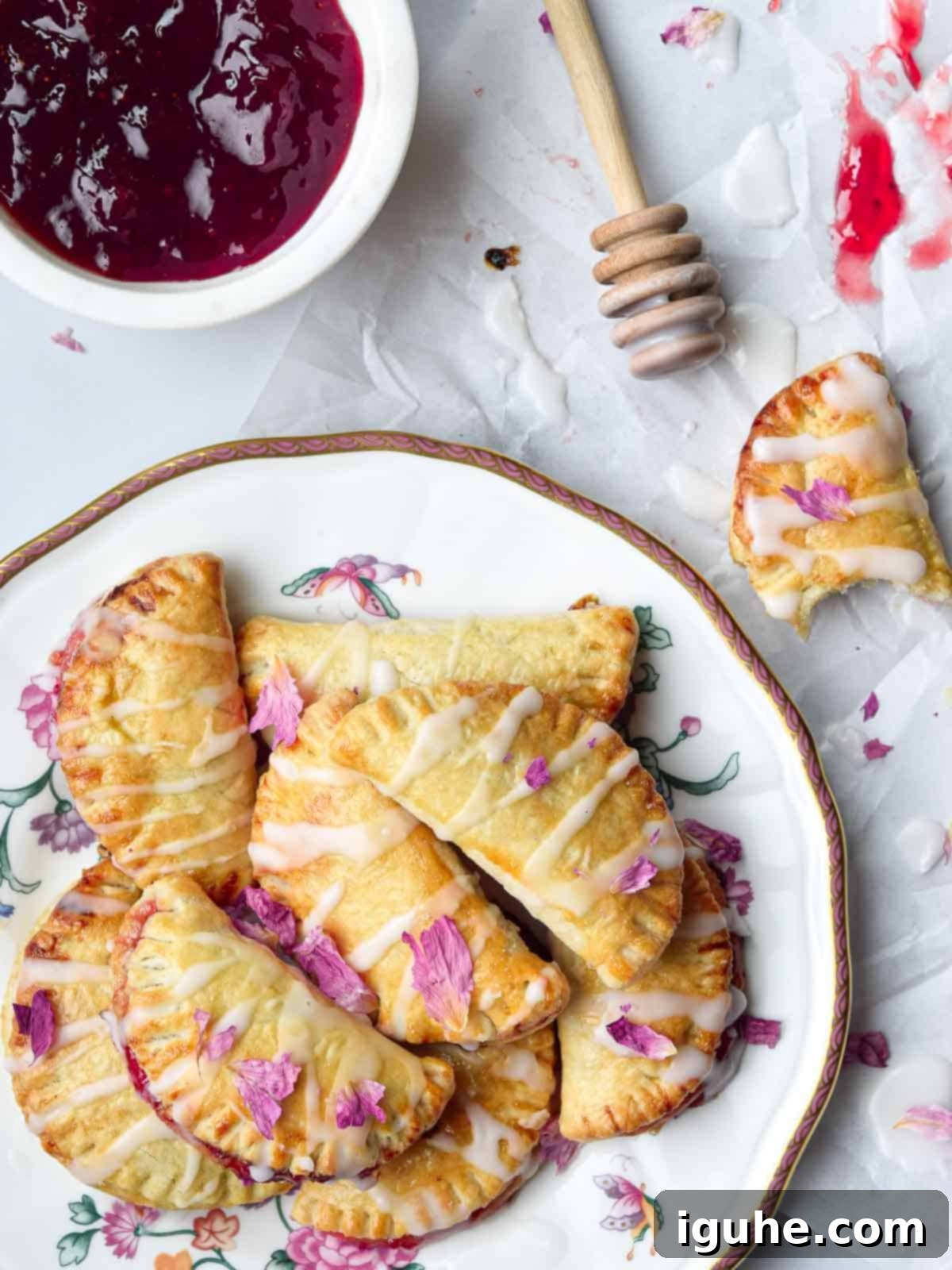 Overhead view of several golden-brown strawberry mini pies arranged on a white plate, with a delicate rose glaze drizzled over them. Fresh rose petals are scattered around, adding an elegant touch.