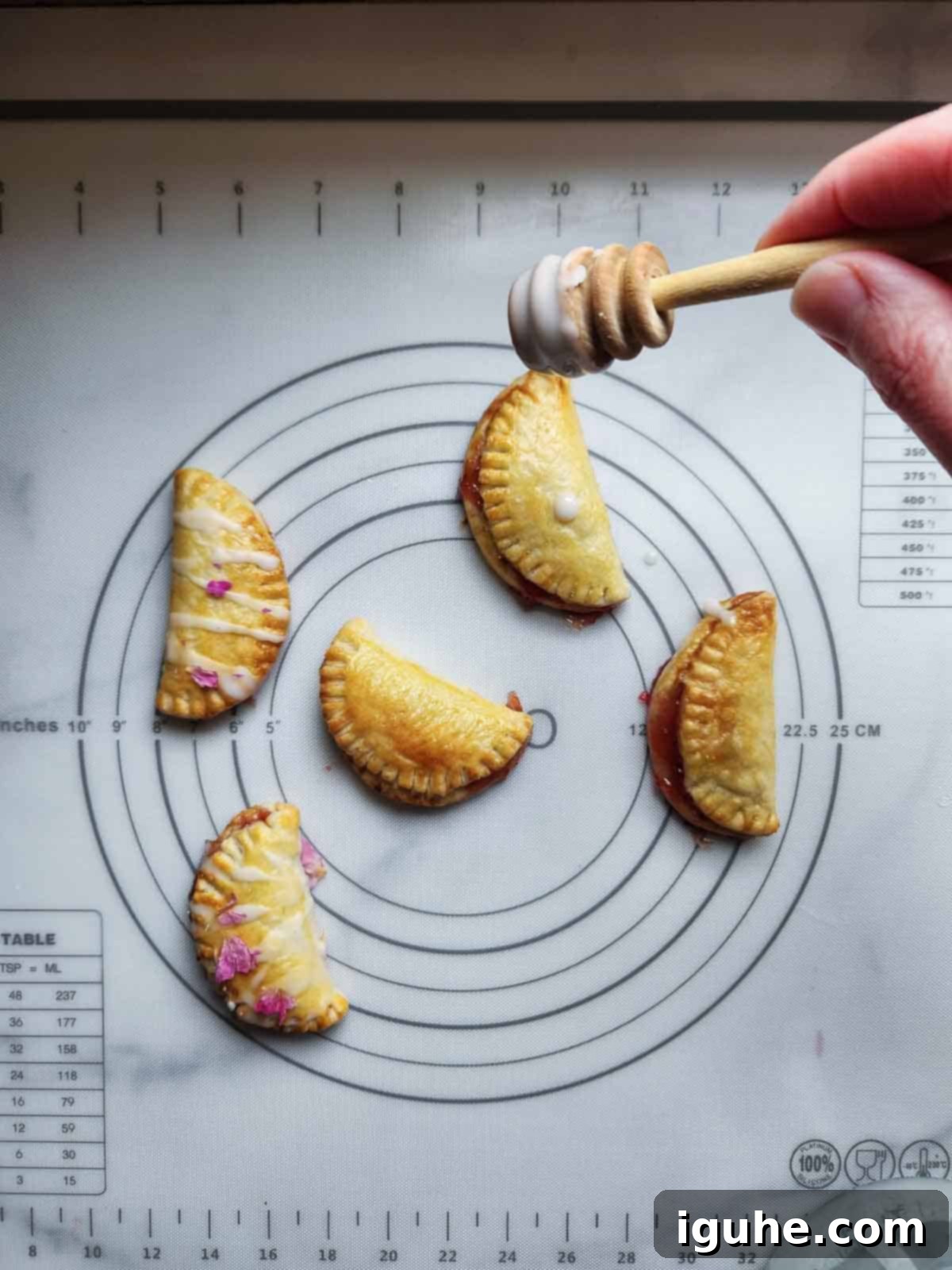 A baker is gently drizzling delicate rose glaze over warm, golden-brown strawberry mini pies arranged on a cooling rack, with a small bowl of extra glaze nearby.