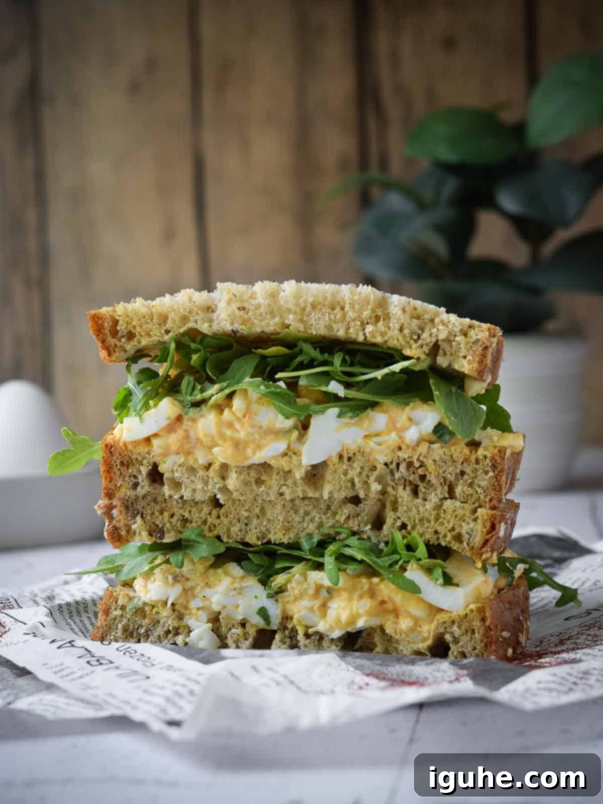 Smoky Paprika and Fresh Chive Egg Salad 3 A perfectly stacked egg salad sandwich with peppery arugula, served on deli paper against a rustic wood background.