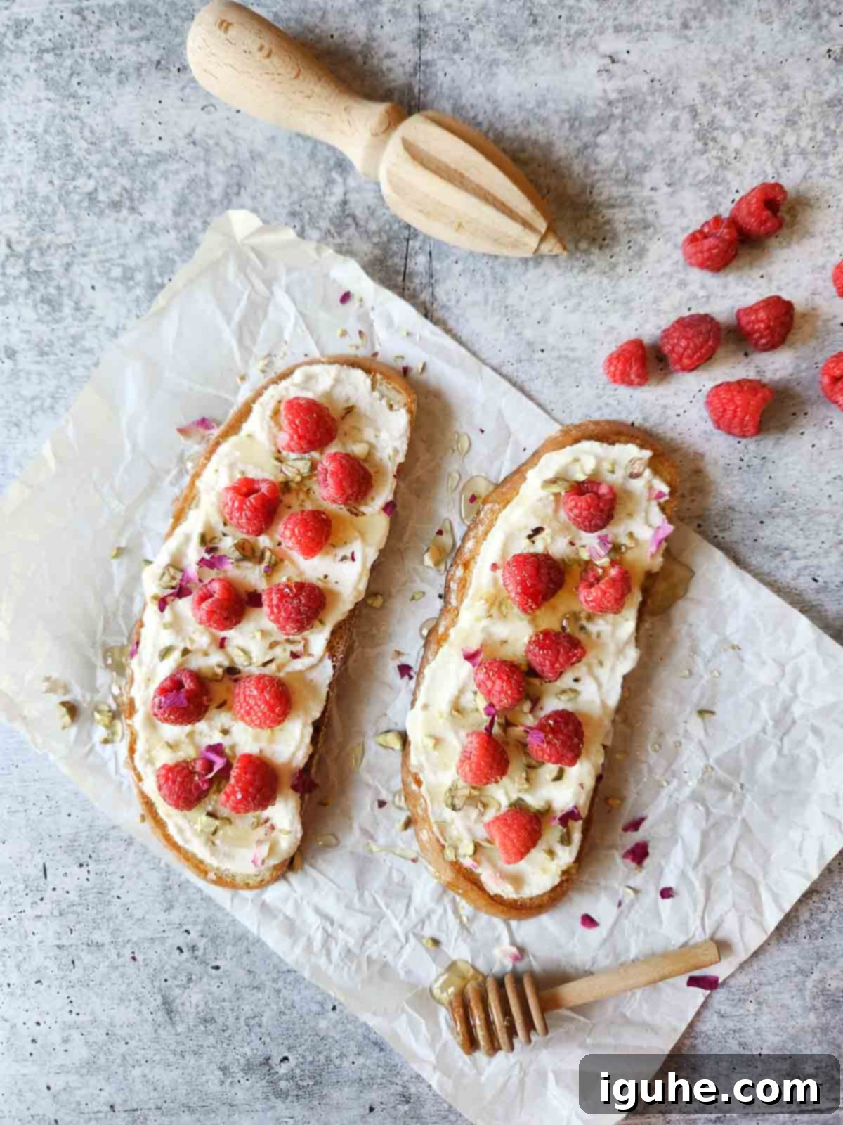 Bright Lemon Raspberry Ricotta Toast 7 Two slices of fully assembled raspberry and ricotta toast on white parchment paper, garnished with pistachios, lemon zest, and honey drizzle.