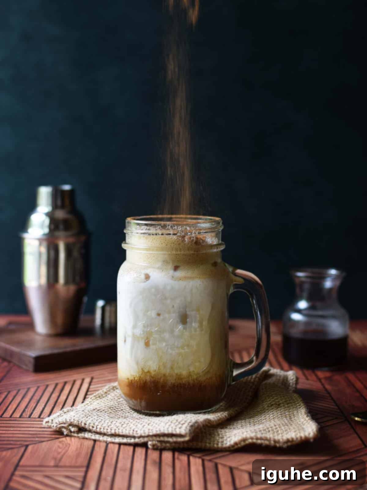 Caramel-Kissed Shaken Chai 9 A hand dusting ground cinnamon over the frothy top of a brown sugar chai iced tea in a clear mug.