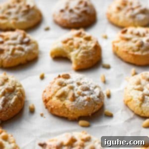 The Easiest Italian Pignoli Cookies 9 Angled view of baked Italian pignoli cookies on a baking sheet covered with white parchment paper. One cookie has a bite taken out.