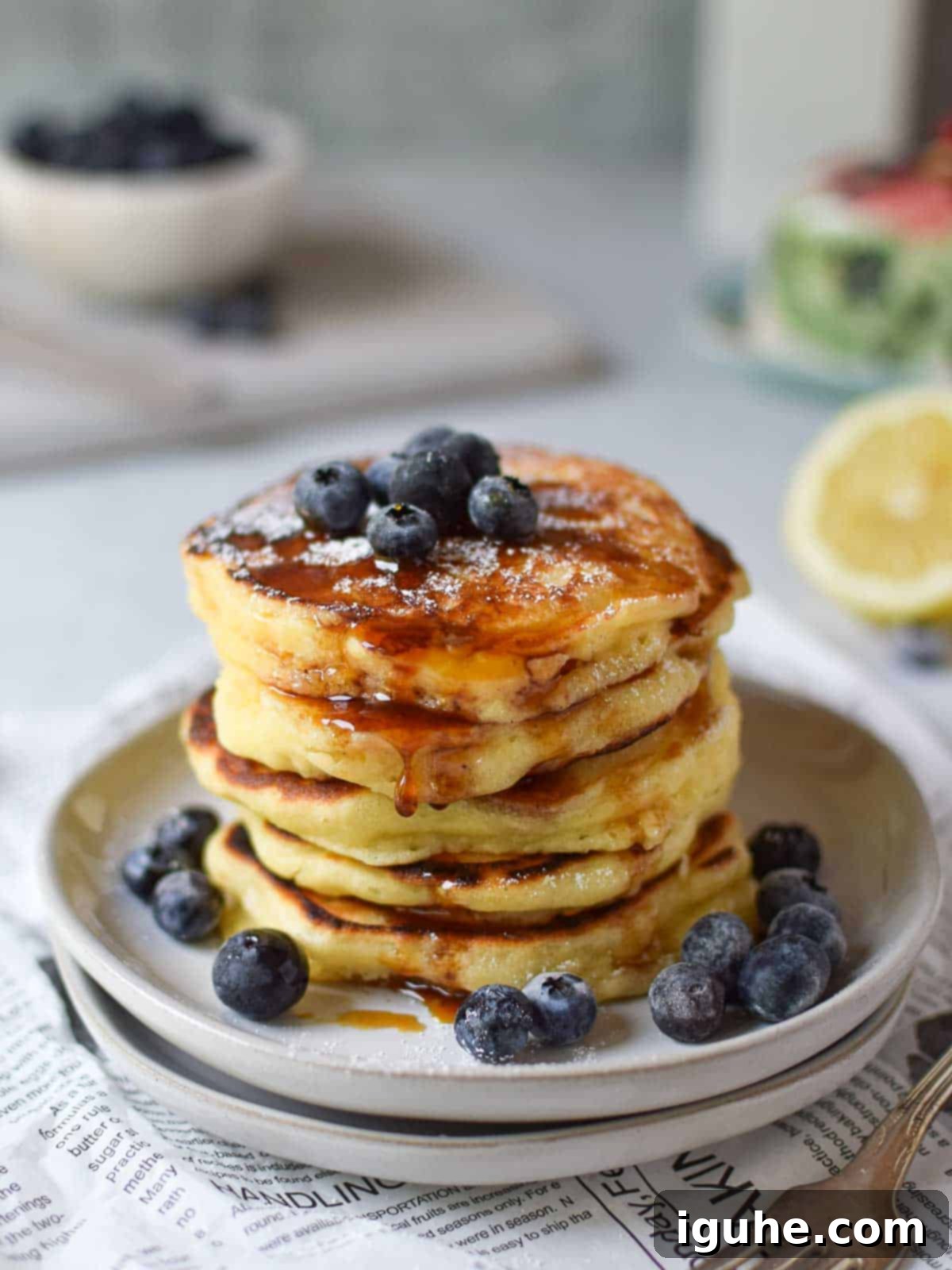 Dreamy Lemon Mascarpone Pancakes 3 Lemon pancakes with maple syrup stacked on plates with blueberries on top and ingredients laid out.