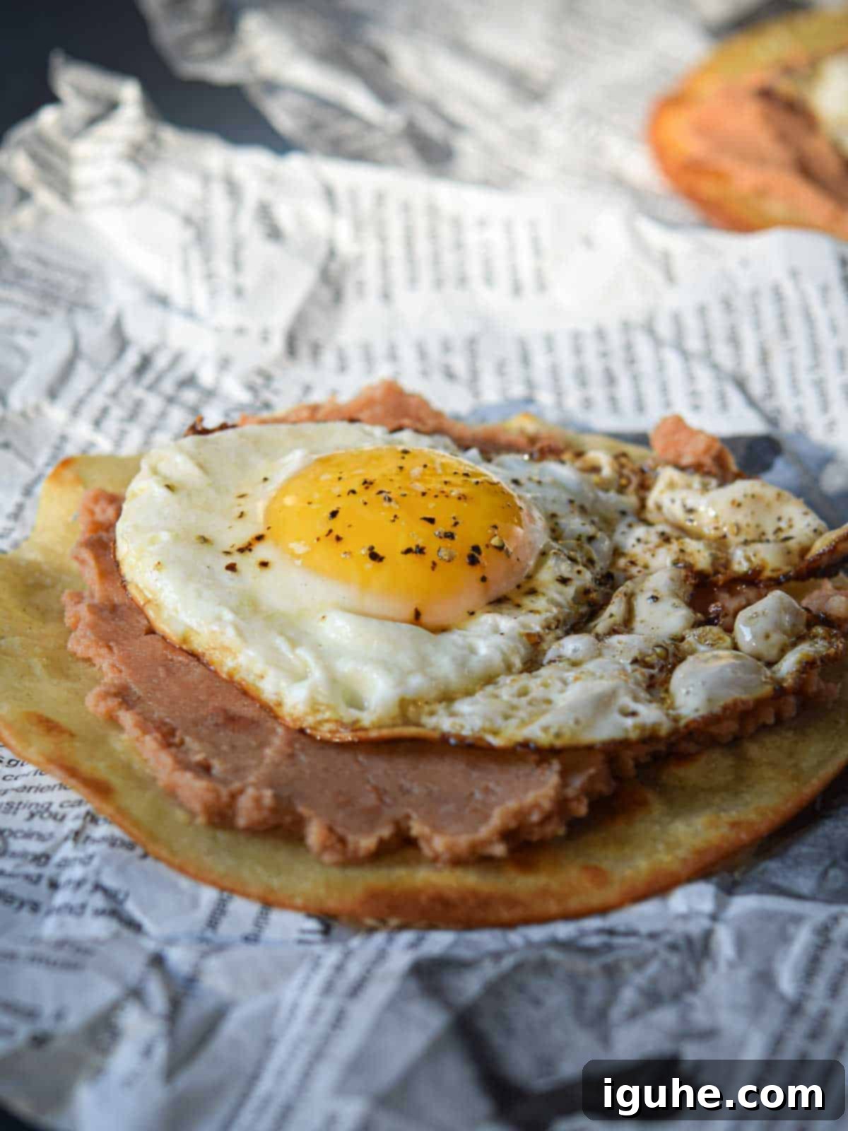 Quick and Delicious Huevos Rancheros Tostadas 7 Fried egg on top of a fried tortilla with refried beans over newspaper.