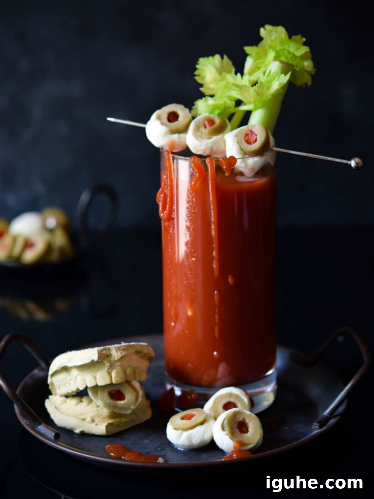 A Halloween Bloody Mary cocktail with a celery stick and two mozzarella and olive 'eyeball' garnishes, set against a dark, moody background. Perfect for spooky season.