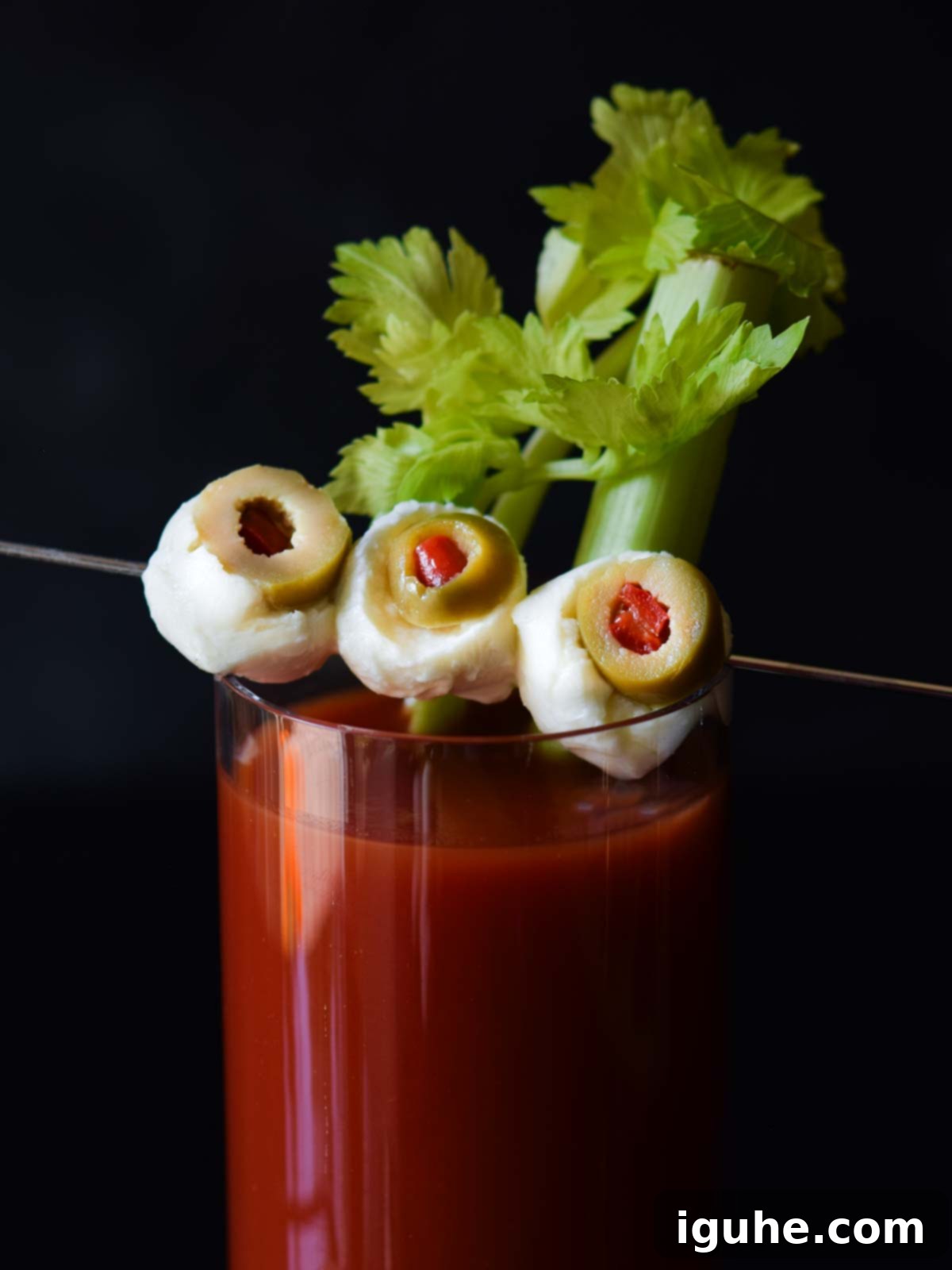 Halloween's Bloody Scary Mary 7 Close-up of three spooky mozzarella and olive 'eyeballs' on a cocktail pick, with the pimento center of the olive creating a bloodshot effect, against a dark background.