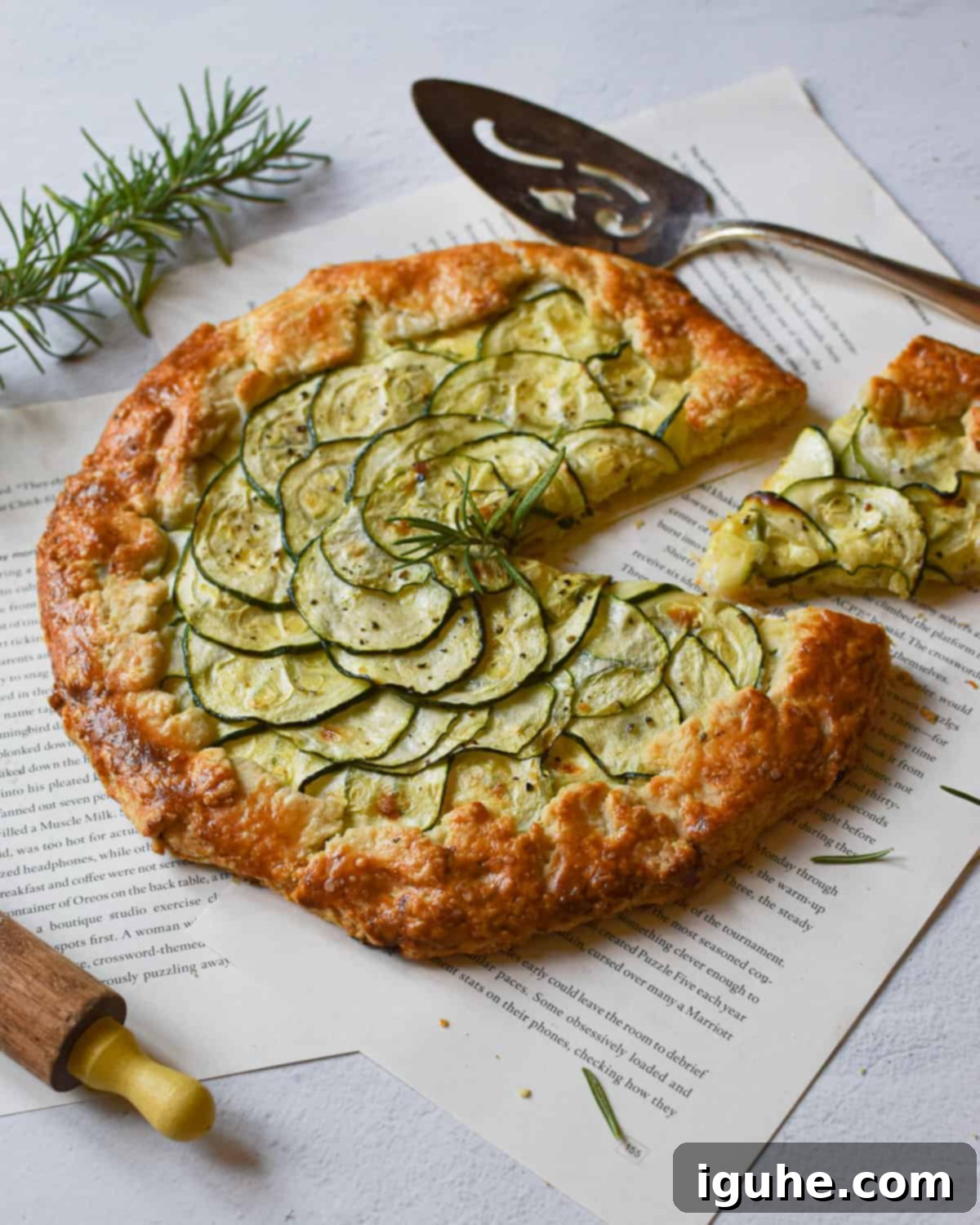 Angled view of zucchini ricotta galette with a slice cut out surrounded by scattered rosemary on a white background.