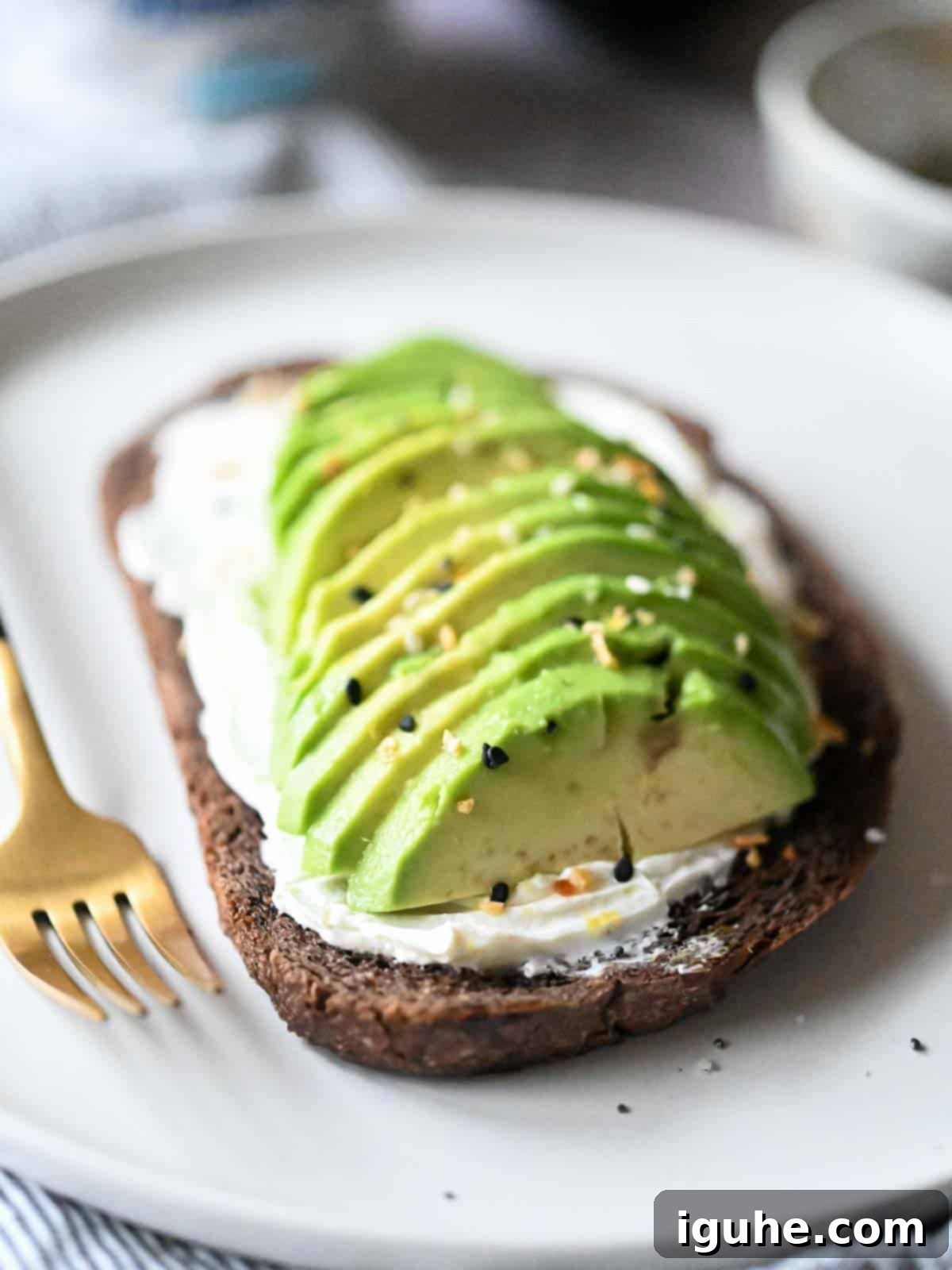 Sliced avocado on pumpernickel bread with cream cheese and spices, garnished with fresh herbs and a hint of lemon.