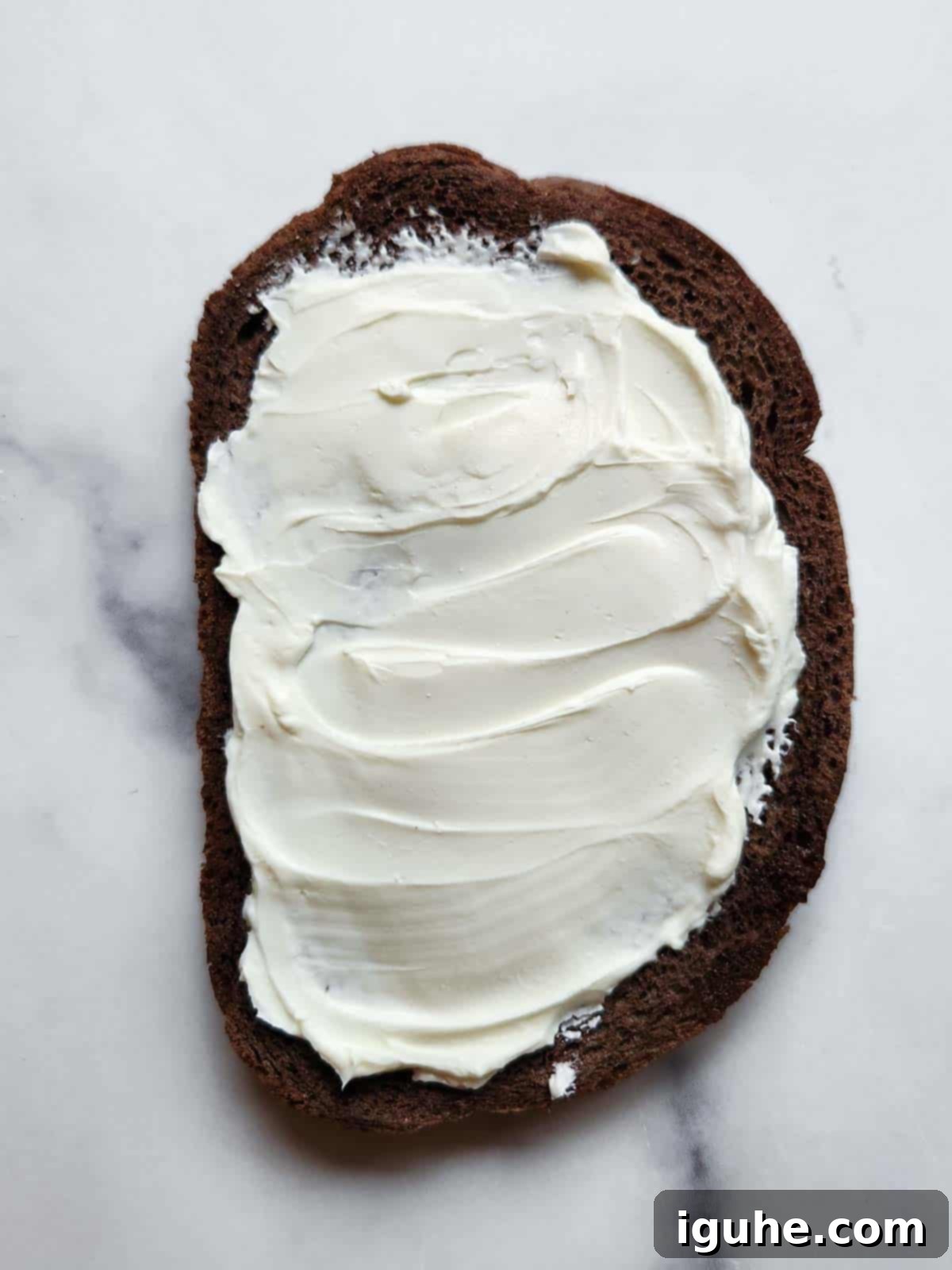 Gourmet Cream Cheese Avocado Toast 5 A slice of toasted pumpernickel bread generously spread with softened cream cheese on a clean white background.