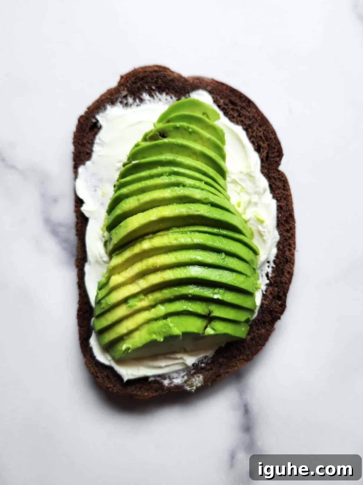 Gourmet Cream Cheese Avocado Toast 6 Toasted pumpernickel bread with cream cheese, topped with neatly sliced avocado, ready for seasoning.