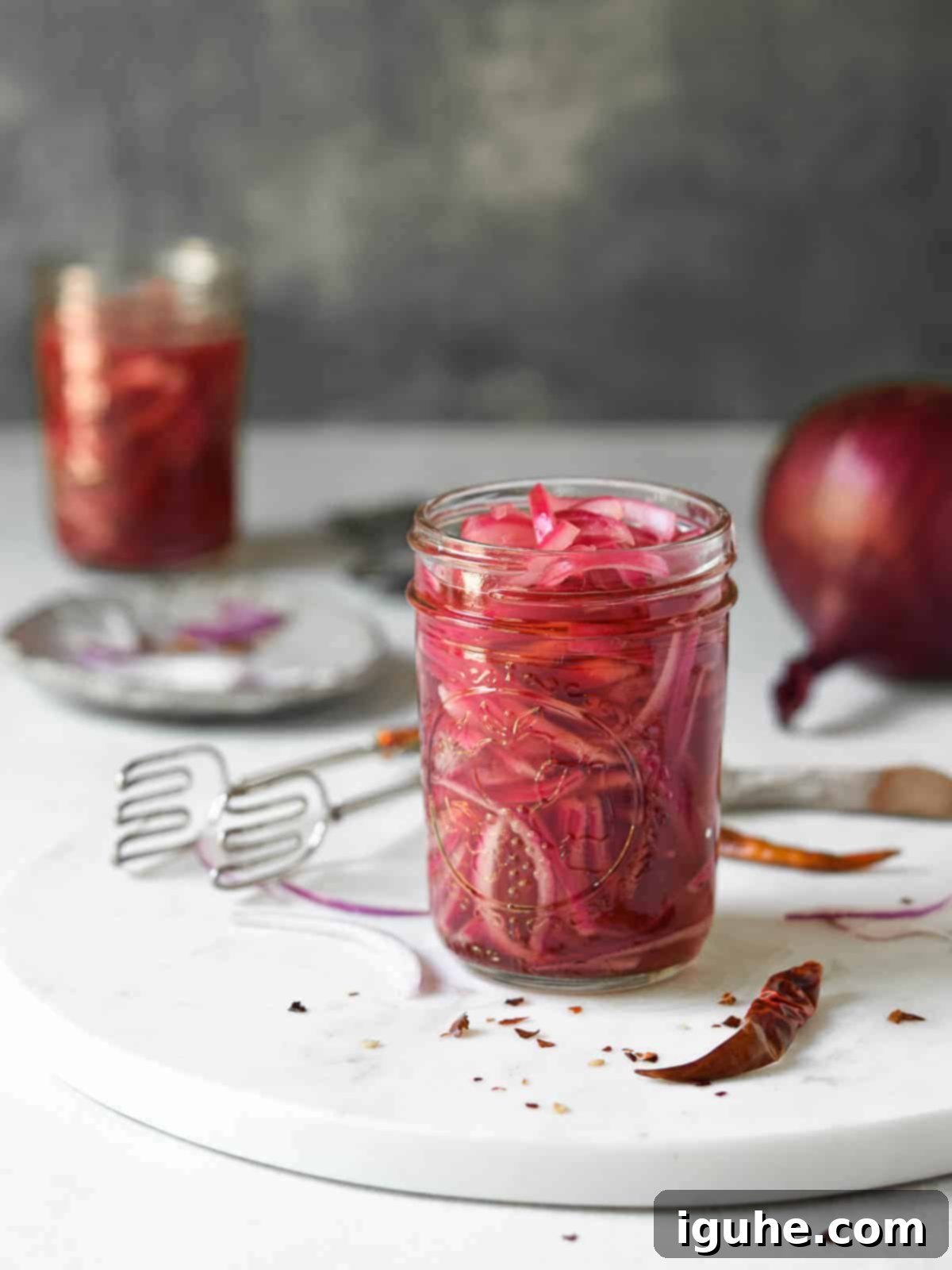 Sweet Heat Pickled Onions 8 A mason jar filled with pickled red onions, surrounded by loose red pepper flakes and allspice on a marble surface.