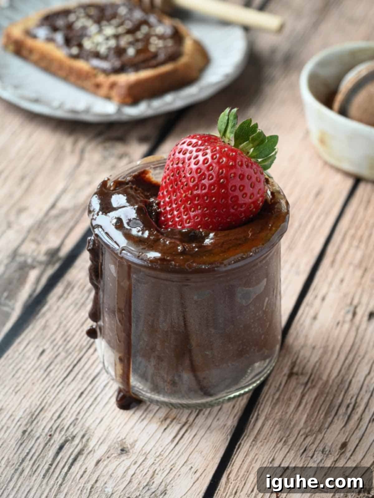 A strawberry dipped into a jar of chocolate tahini spread on wood.