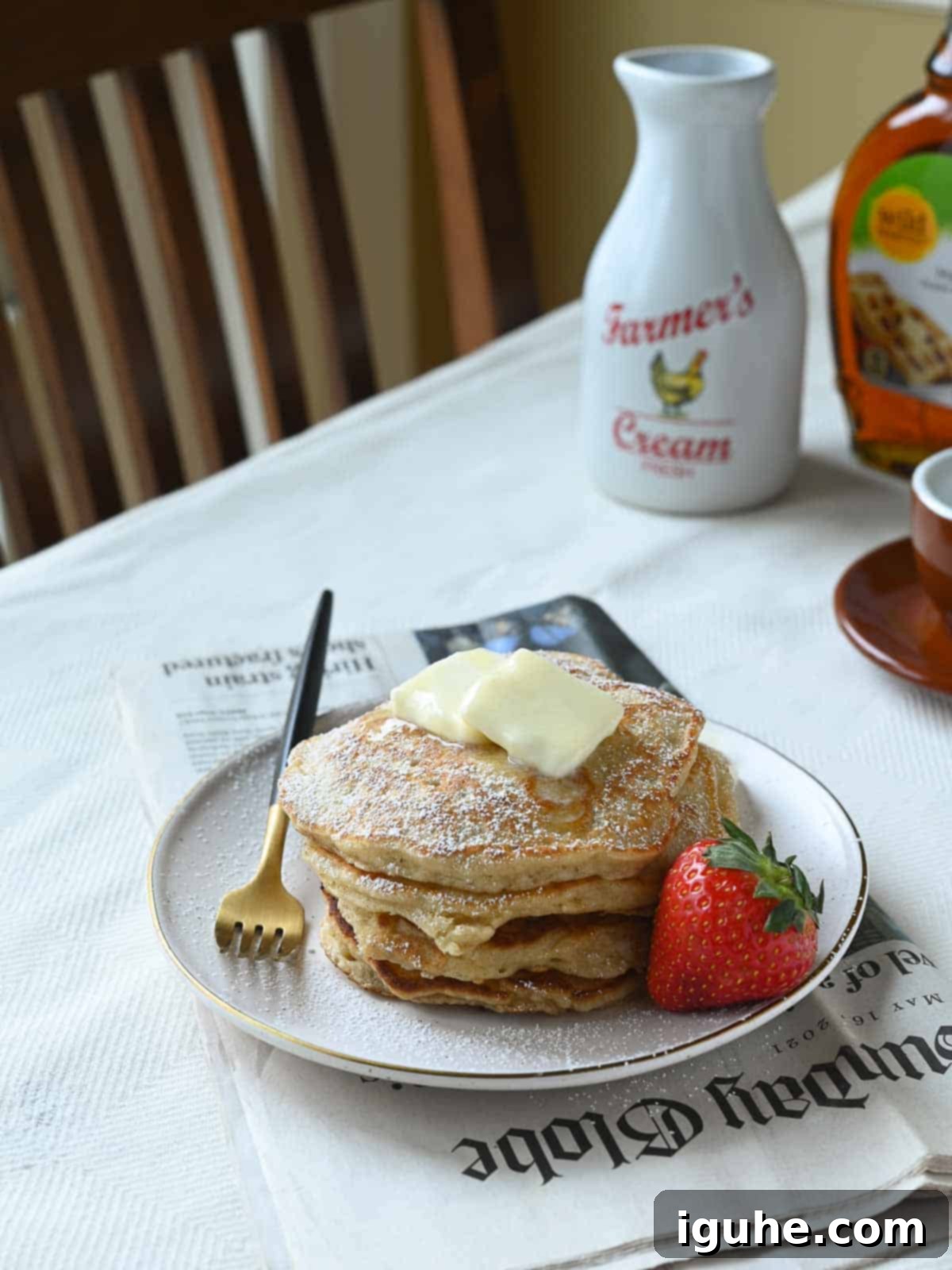 The Art of Perfectly Puffed Pancakes 3 Pancakes on a kitchen table with a strawberry to the side and the morning paper under the plate.