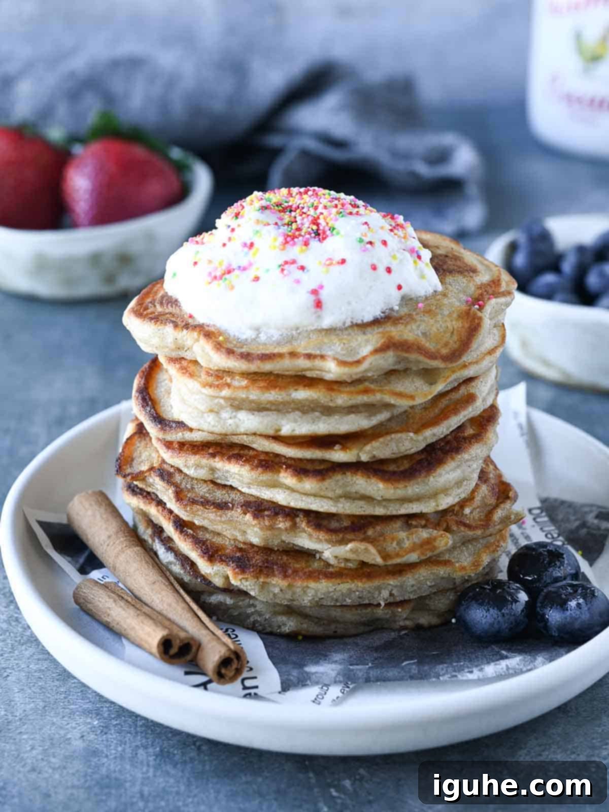 A stack of golden-brown cinnamon sweet cream pancakes with whipped cream, maple syrup, and festive sprinkles on top, resting on a clean grey surface, ready to be enjoyed.