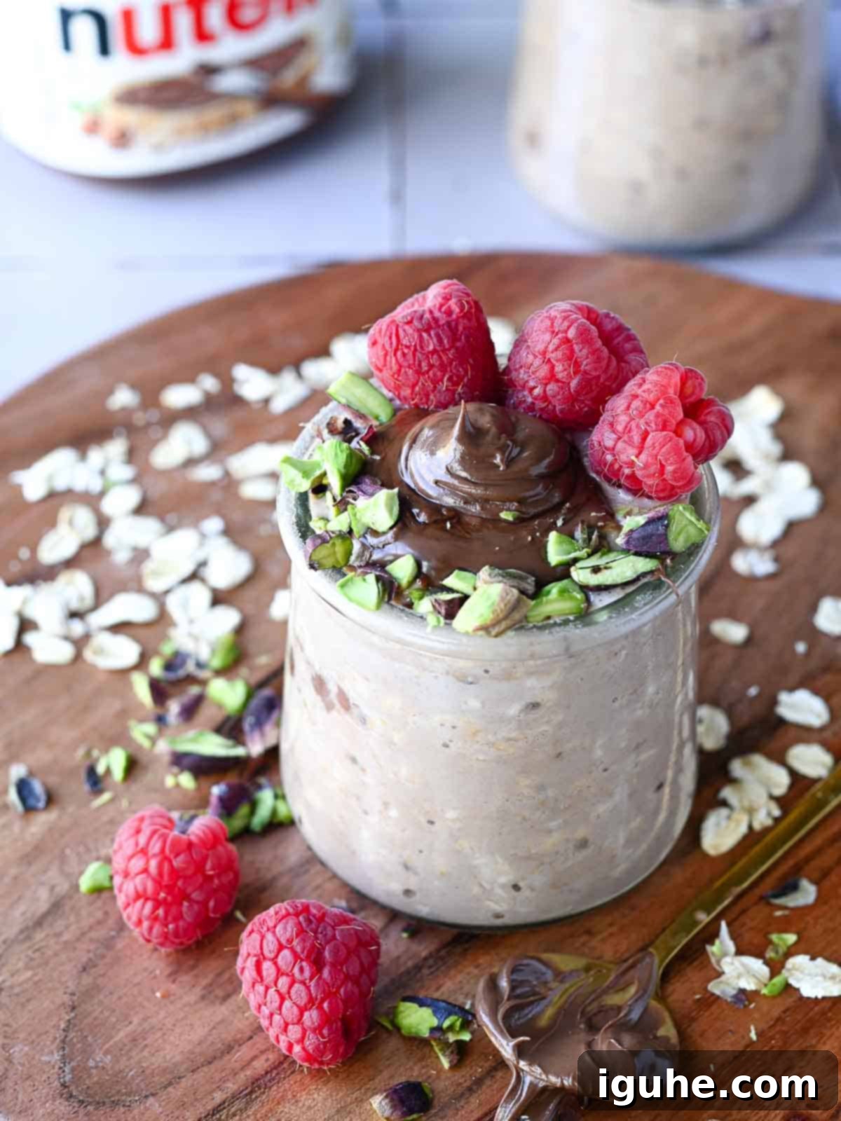 Decadent Nutella Overnight Oats 6 Nutella overnight oats in a clear jar with raspberries and pistachios on wood.