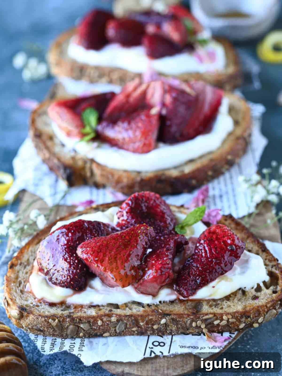 Three slices of labneh toast with strawberries on a blue surface, garnished with mint and rose petals.