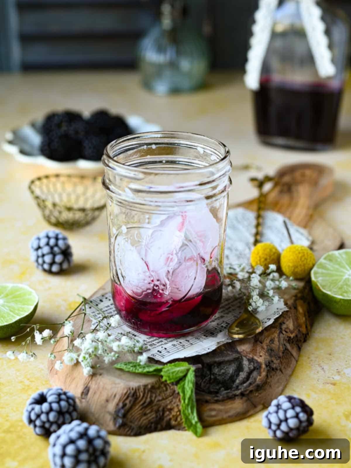 Blackberry Sparkler 3 Ice and Blackberry syrup in a clear glass on top of a yellow surface with blackberries around it.