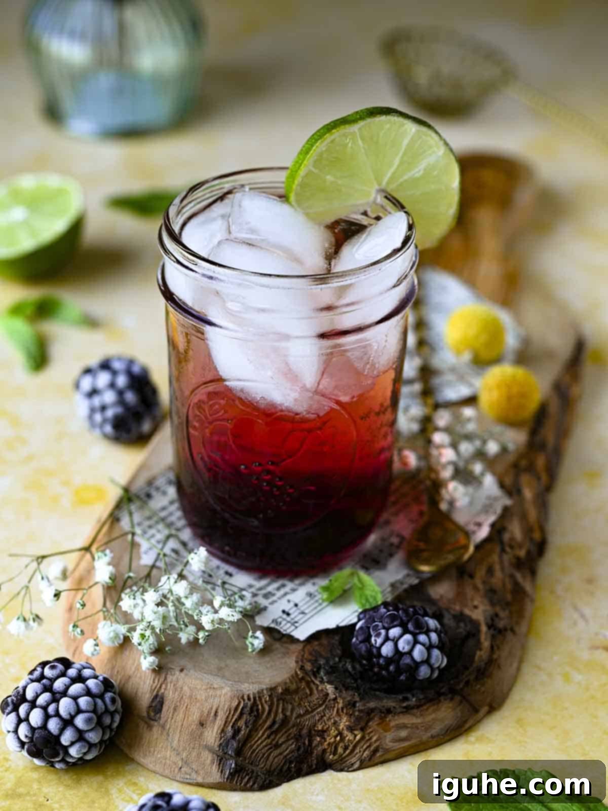 Blackberry Sparkler 5 Blackberry soda in a clear glass on wood with blackberries and flowers around it.