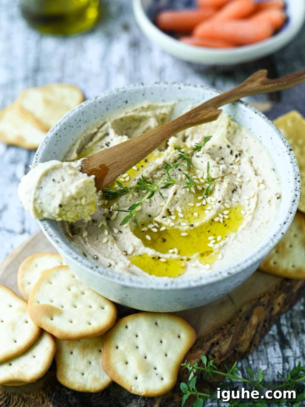 Silken Butter Bean Hummus 8 Hummus with a wooden spreader in a white bowl on a blue surface.