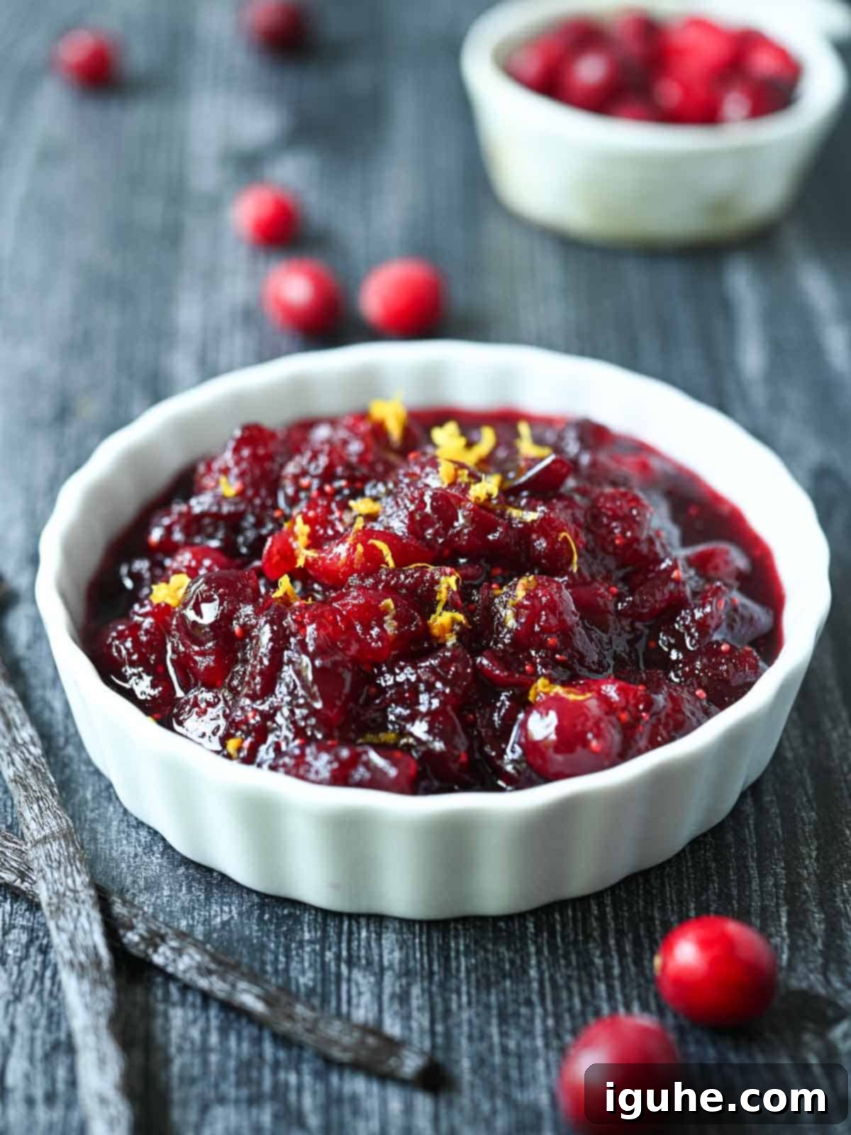 Cranberry compote in a white ramekin with cranberries and vanilla beans beside it, showcasing its rich texture and vibrant color.