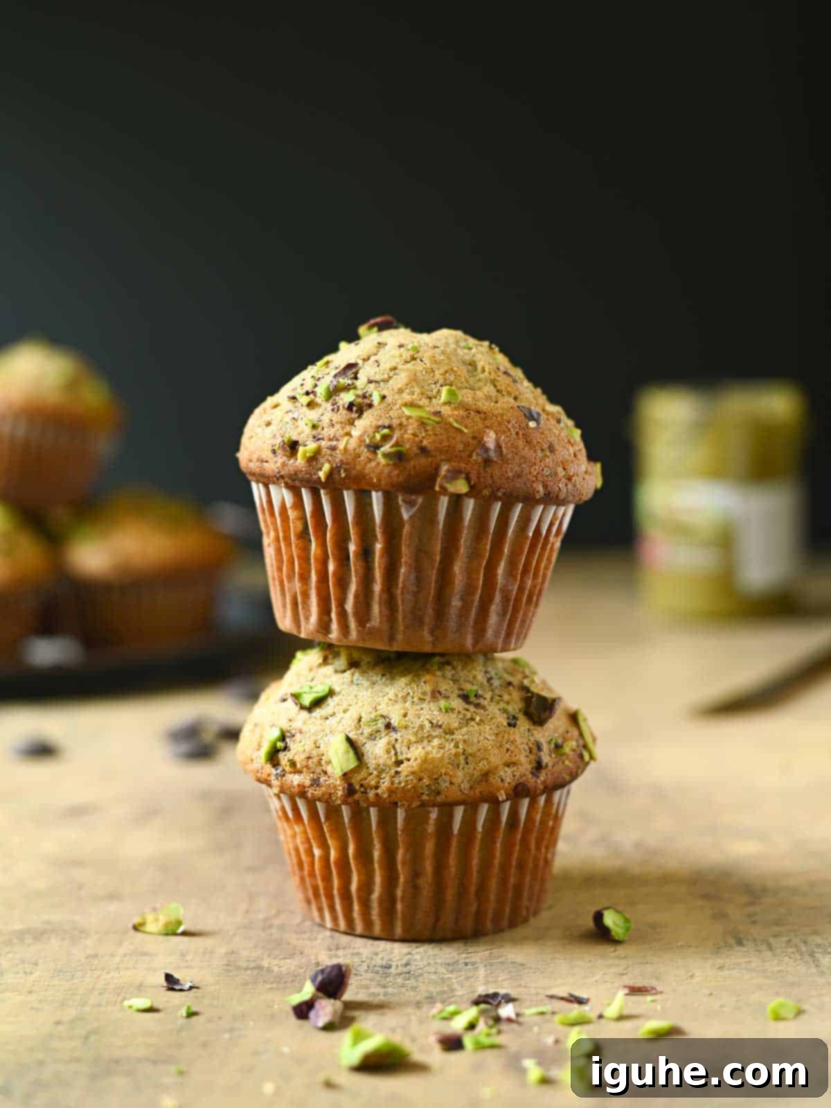 Decadent Pistachio Chocolate Chip Muffins 6 Two freshly baked pistachio muffins are elegantly stacked on a rustic brown surface, showcasing their golden-brown tops adorned with crushed pistachios and coarse sugar, with the dark chocolate peeking through.