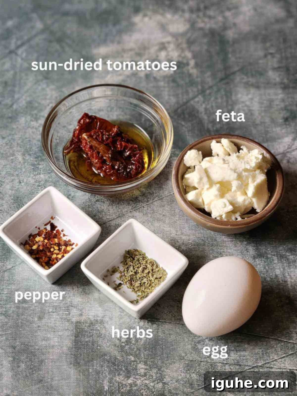 Sun-Kissed Feta and Tomato Fried Eggs 3 Assortment of fresh ingredients for sun-dried tomato feta fried eggs, including eggs, a block of feta cheese, sun-dried tomatoes in oil, and sprigs of fresh herbs, laid out on a grey kitchen surface.