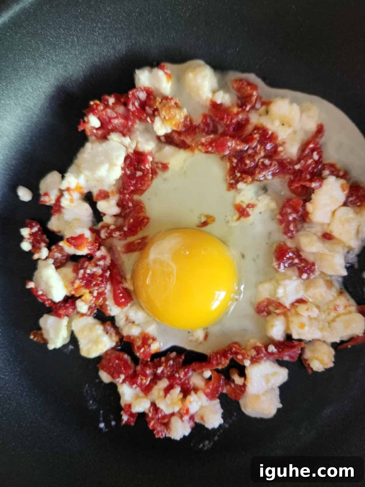 Sun-Kissed Feta and Tomato Fried Eggs 5 A raw egg perfectly cracked into the center of the crisping feta and sun-dried tomato ring in a hot skillet, ready for cooking.