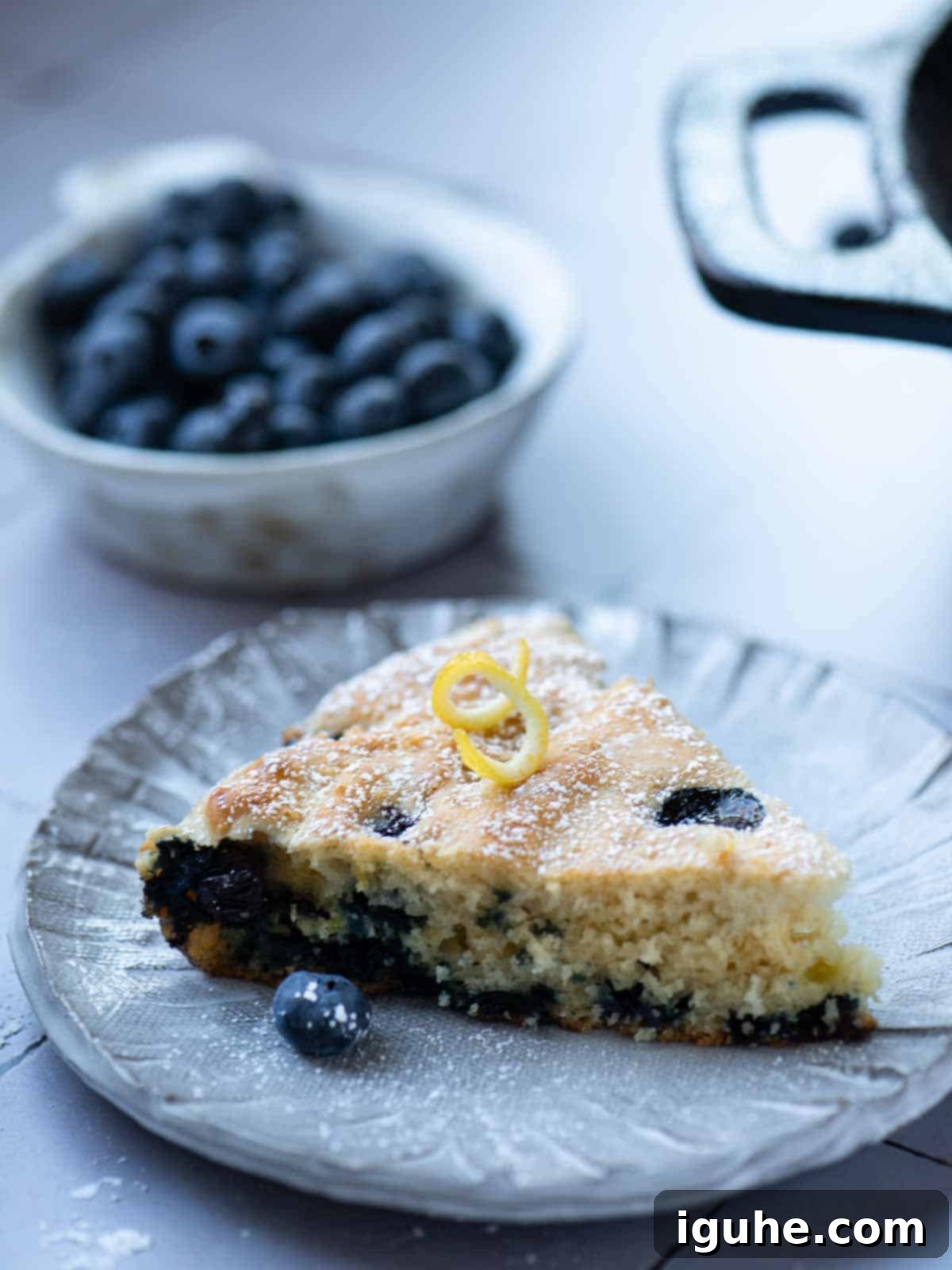 Blueberry Burst Skillet Pancake 10 A slice of blueberry skillet pancake on a grey plate with blueberries in the background.
