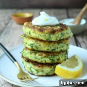 Crispy Zucchini Parmesan Fritters 10 Stacked crispy Parmesan zucchini pancakes on a beige plate, garnished with lemon and fresh herbs, with a fork on the side.